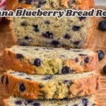 Sliced banana blueberry bread on a wooden board, showing a moist, golden loaf filled with juicy blueberries. A few fresh blueberries are scattered around, and a red-striped towel is visible in the corner. The image features the text "Banana Blueberry Bread Recipe" across the top.