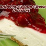 Slice of cranberry cream cheese dessert with a graham cracker crust, creamy cheese layer, and glossy cranberry topping, served on a green plate with a fork and the text "Cranberry Cream Cheese Dessert"